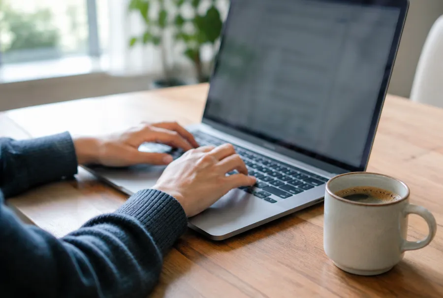 Hände auf der Tastatur eines Laptops auf einem Holztisch neben einer Tasse Kaffee, der Bildschirm ist unscharf